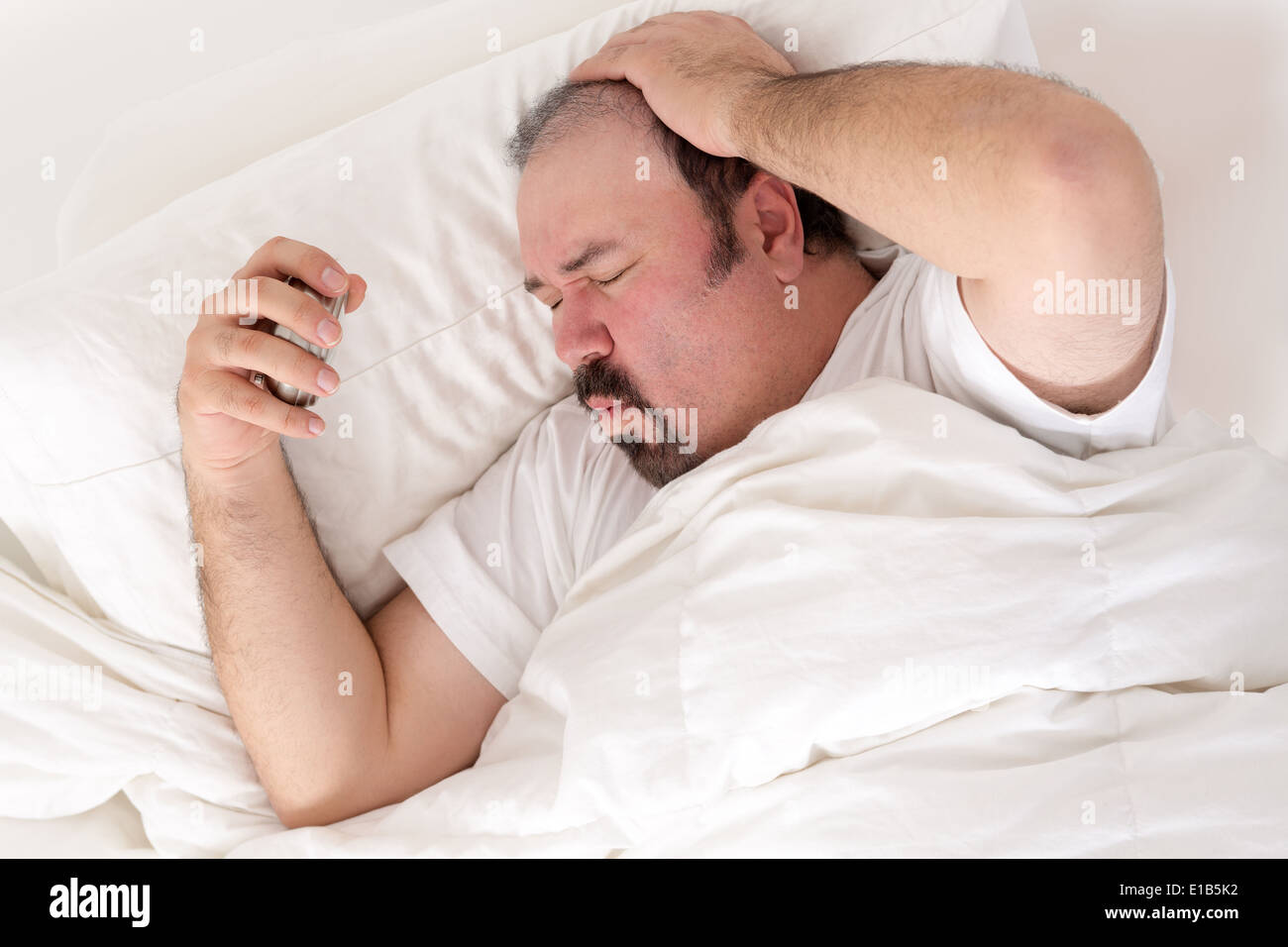 Man suffering from a hangover clutching his head in one hand and alarm clock in the other as he suffers in the morning wondering Stock Photo