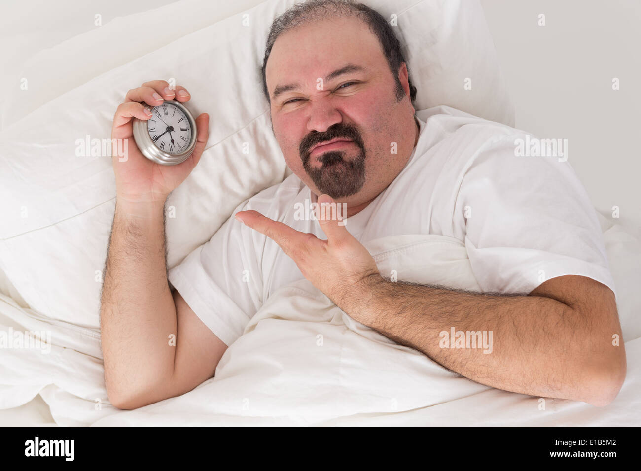 Middle-aged bearded man kept awake by noisy neighbors lying in bed grimacing and pointing to the time on his alarm clock Stock Photo
