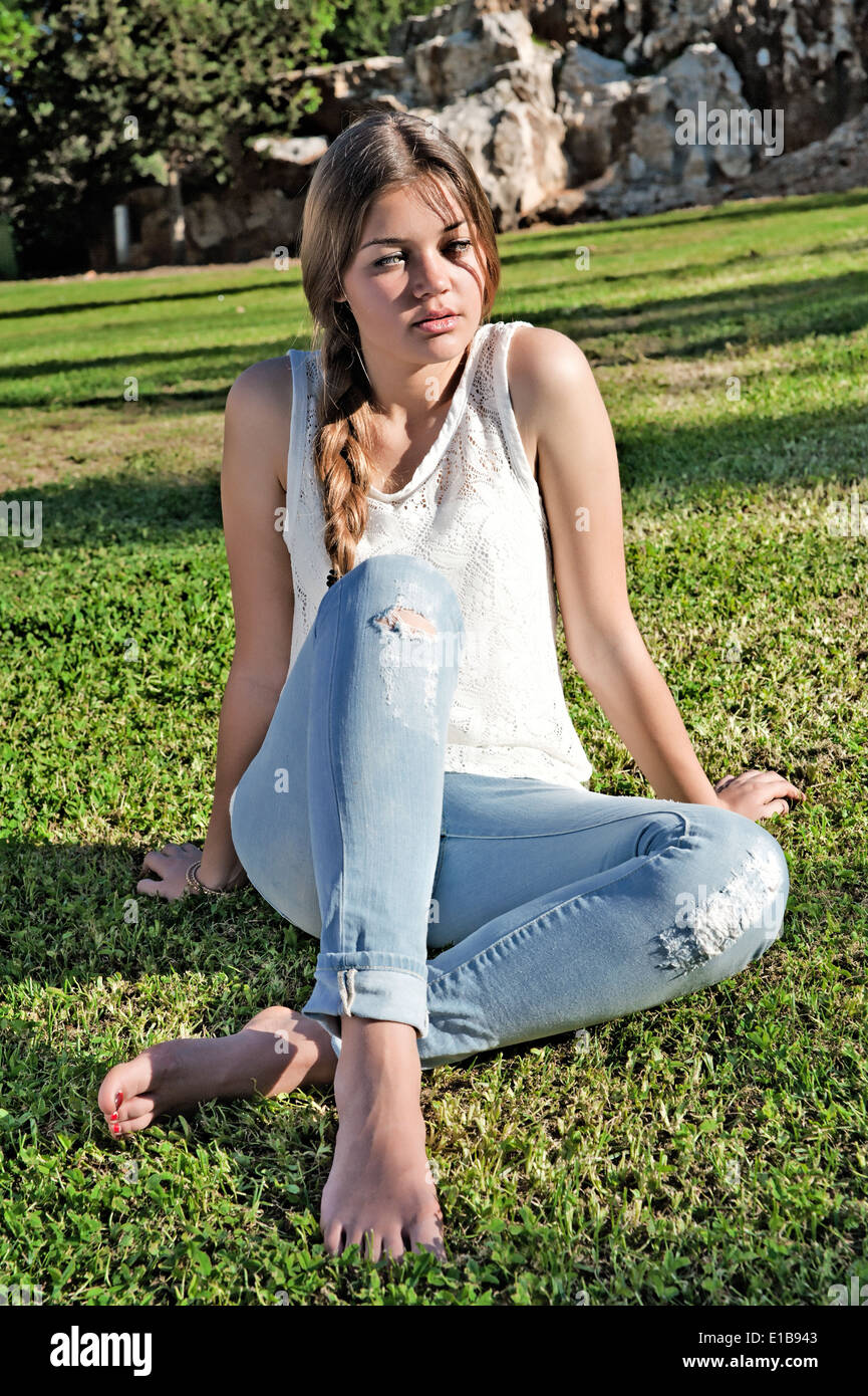 Barefoot girl in ripped jeans sitting on the grass in the park against the rocks Stock Photo