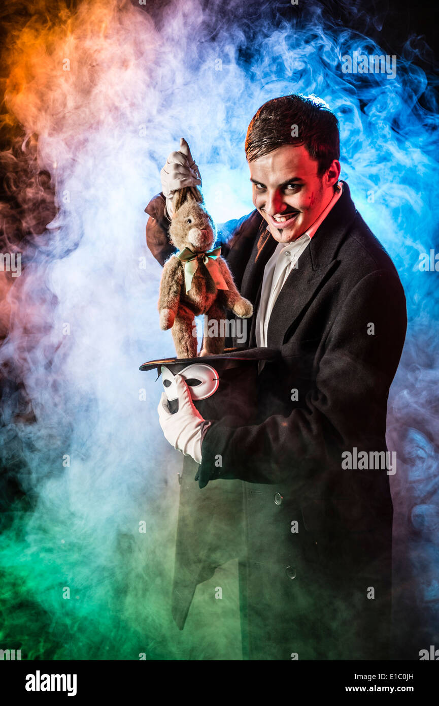 An evil sinister bad magician wreathed in smoke pulling a child's soft toy rabbit out of a top hat Stock Photo