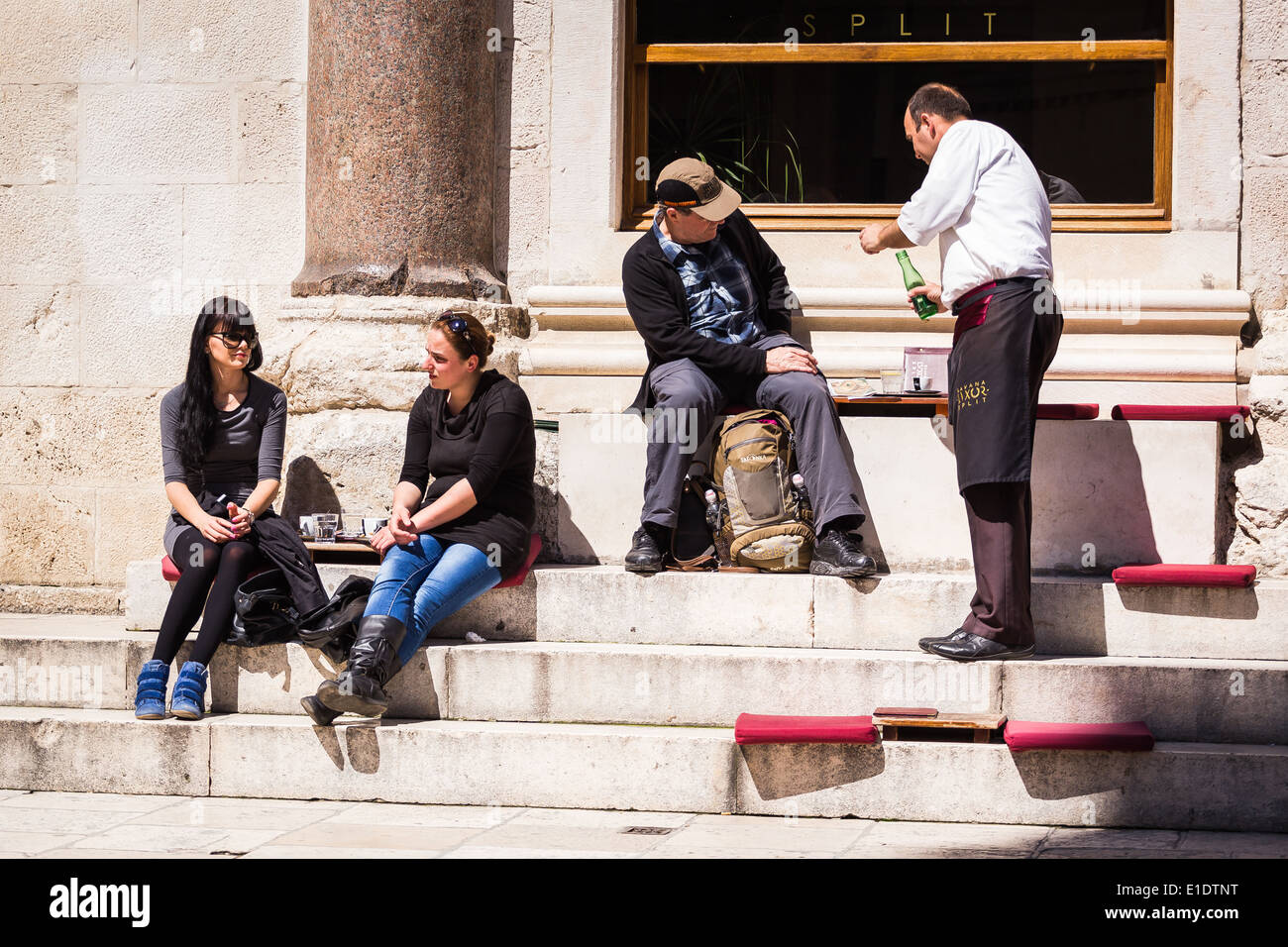 Two women sit enjoying drinks and conversation outside the Luxor hotel in Peristil, Split Croatia as a waiter serves a man Stock Photo