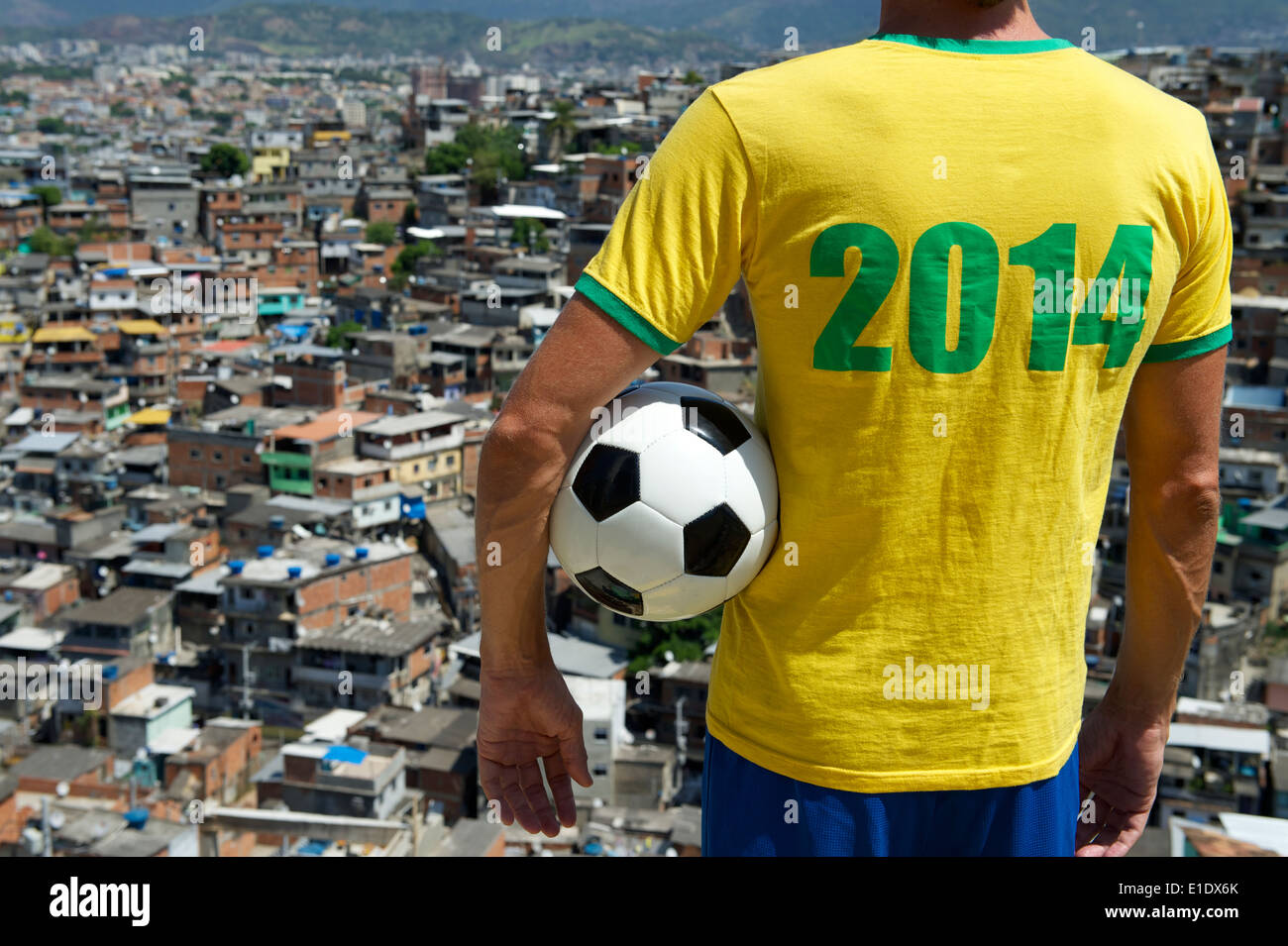Brazilian football player standing in 2014 shirt holding soccer ball in front of favela hillside slum background in Rio Brazil Stock Photo
