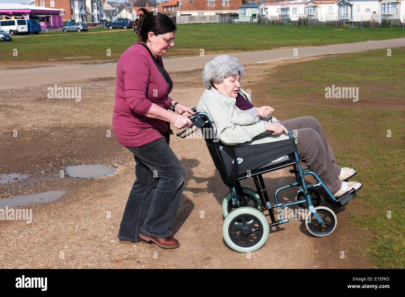 Elderly lady in wheelchair being wheeled across rough ground by her carer or assistant. Stock Photo