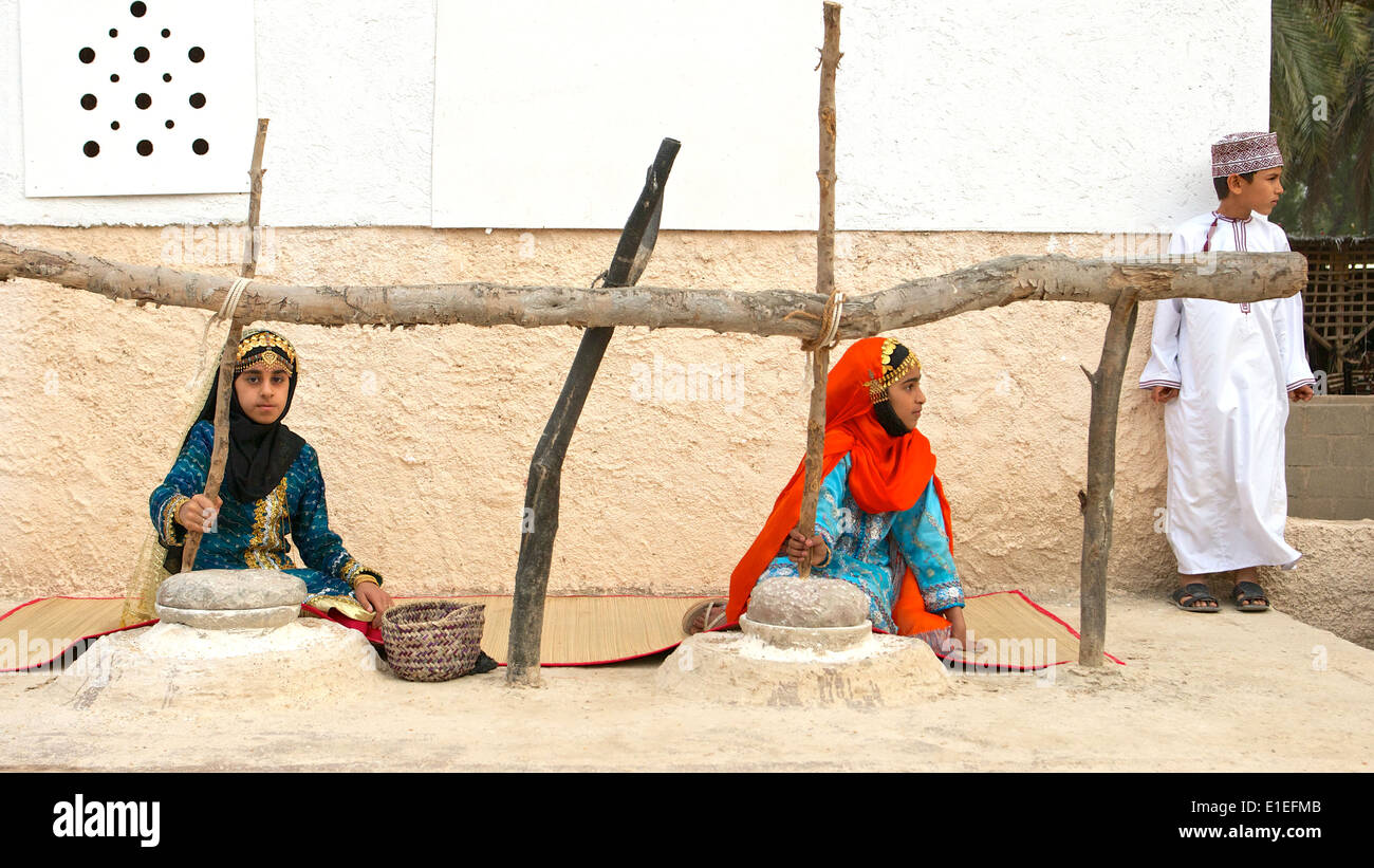 Two Omani girls in traditional dress grinding corn, while their brother waits in the background, in Muscat, Sutanate of Oman. Stock Photo