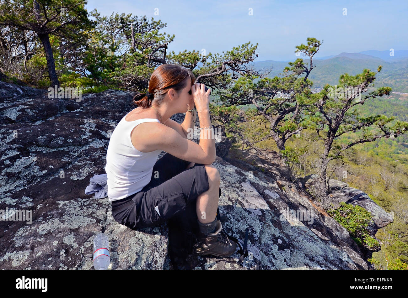 A woman hiker on a rock overlook using her binoculars. Cell phone on her pocket. Stock Photo