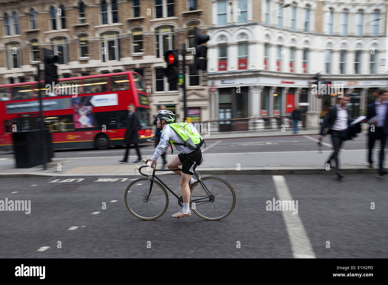 Man wearing shorts, helmet and i-viz yellow backpack rides his racing road bicycle along a busyLondon street. Stock Photo