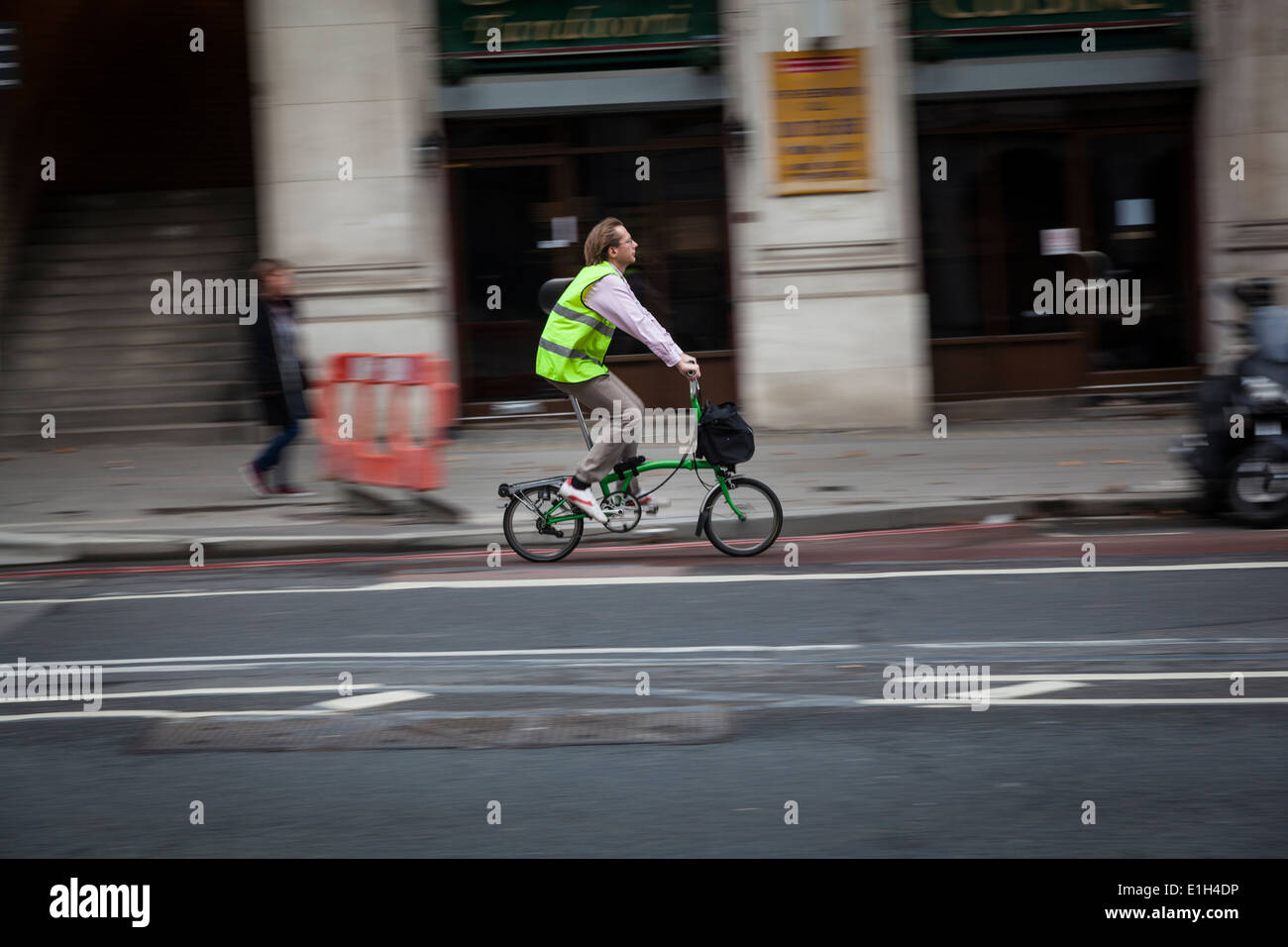 Man wearing a yellow hi-viz jacket rides his Brompton collapsible (folding) bicycle,  with a front carrybag. Stock Photo