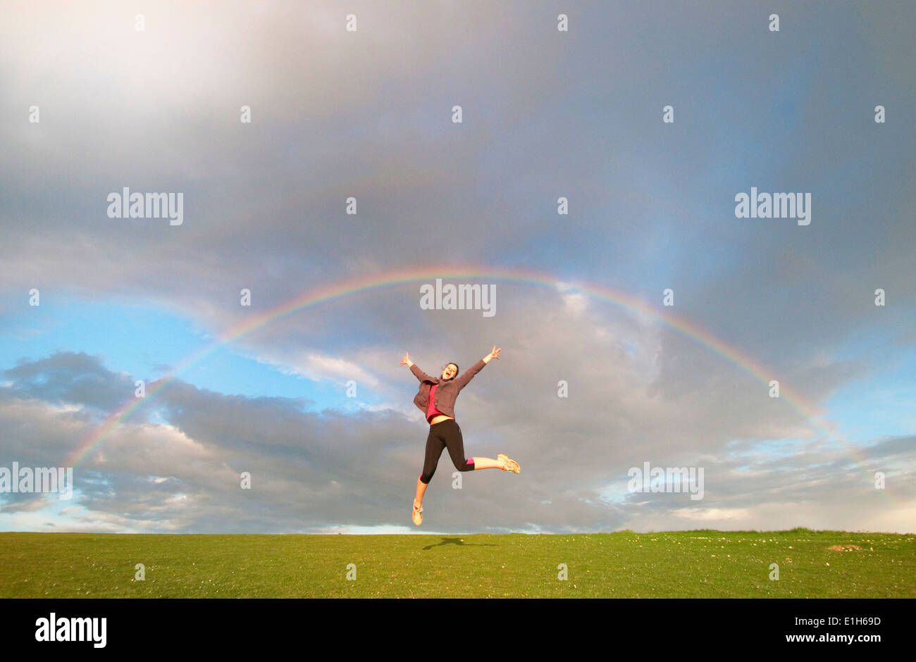 Young female hiker jumping mid air below rainbow Stock Photo