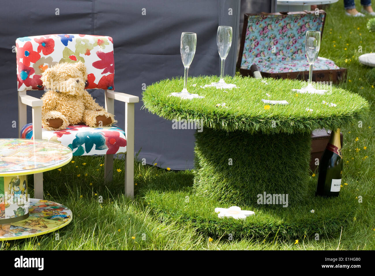 Teddy bear sitting on a chair beside a Astroturf table and wine glasses Stock Photo