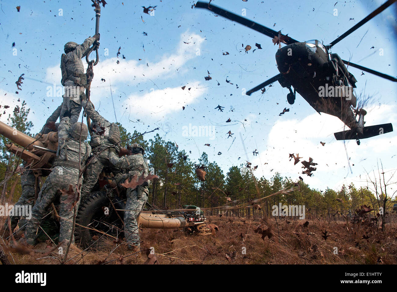 Leaves and twigs are whipped into the air by the rotor wash of UH-60 Black Hawk helicopter as soldiers prepare to hook up an M1 Stock Photo