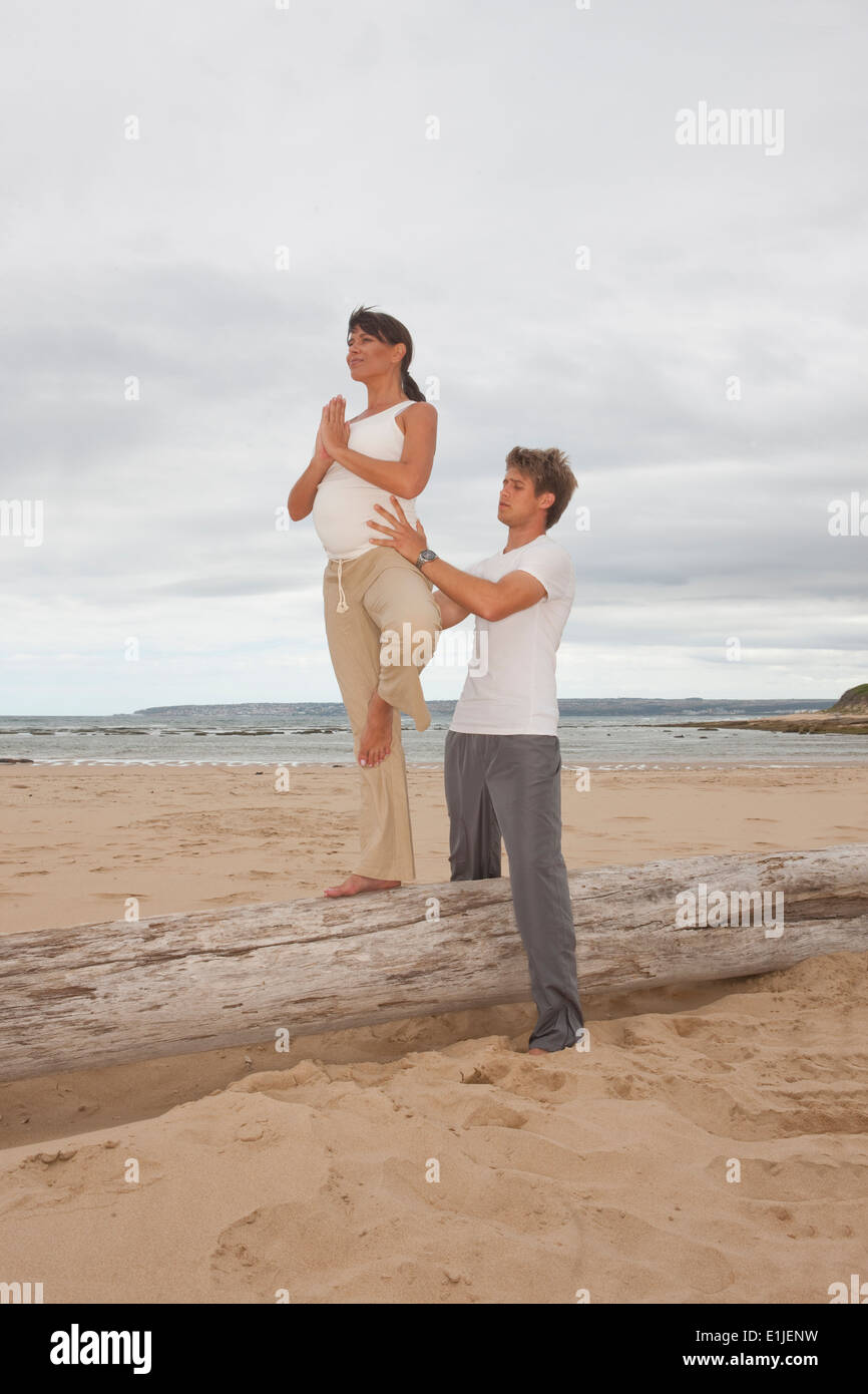 Pregnant mid adult woman with personal trainer practicing yoga at beach Stock Photo