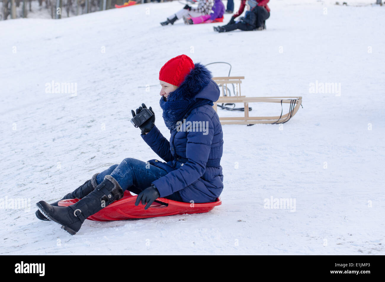 young woman sit on plastic sledge with blue feather jacket and camera in hand Stock Photo