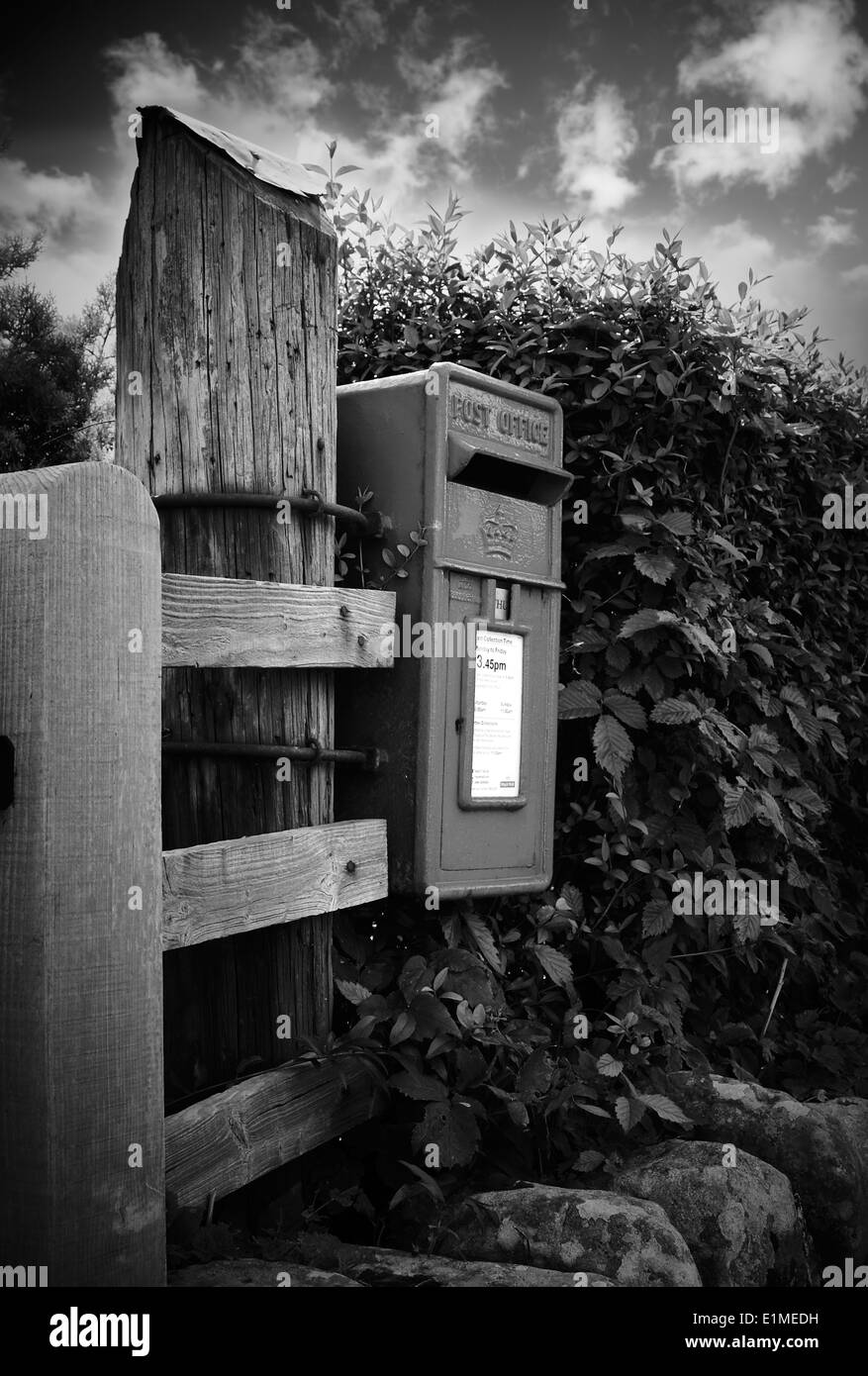 Small royal mail post box attached to a large wooden post in rural setting. Black & white image with dramatic sky Stock Photo