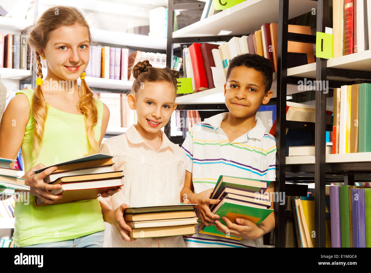 Three friends with books standing near bookshelf Stock Photo