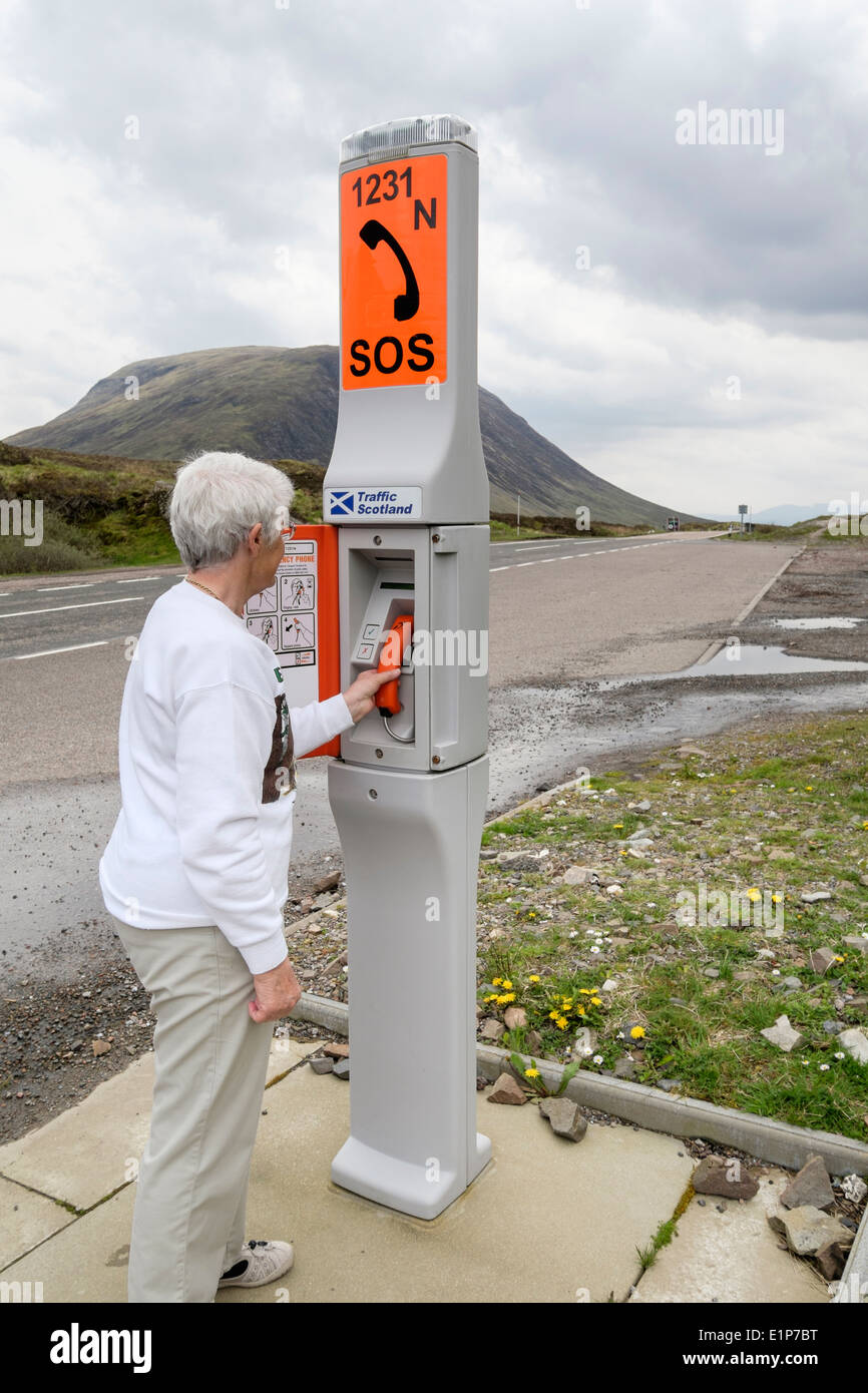 Senior woman using an emergency SOS telephone beside a remote country road calling for roadside assistance help. Glen Coe Highland Scotland UK Britain Stock Photo