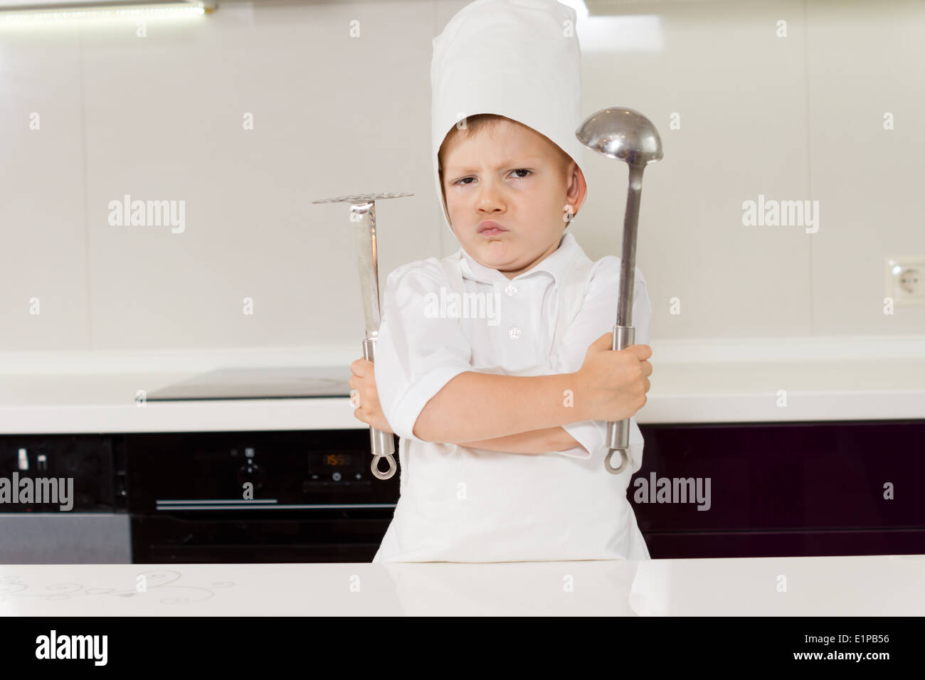 Young boy chef protecting his secret recipe standing gripping his utensils with folded arms and a pugnacious expression Stock Photo