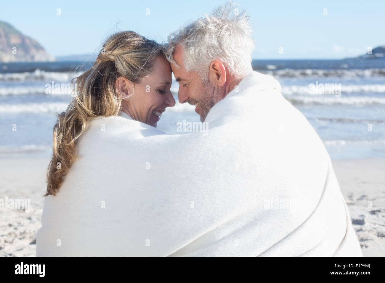 Couple sitting on the beach under blanket smiling at each other Stock Photo