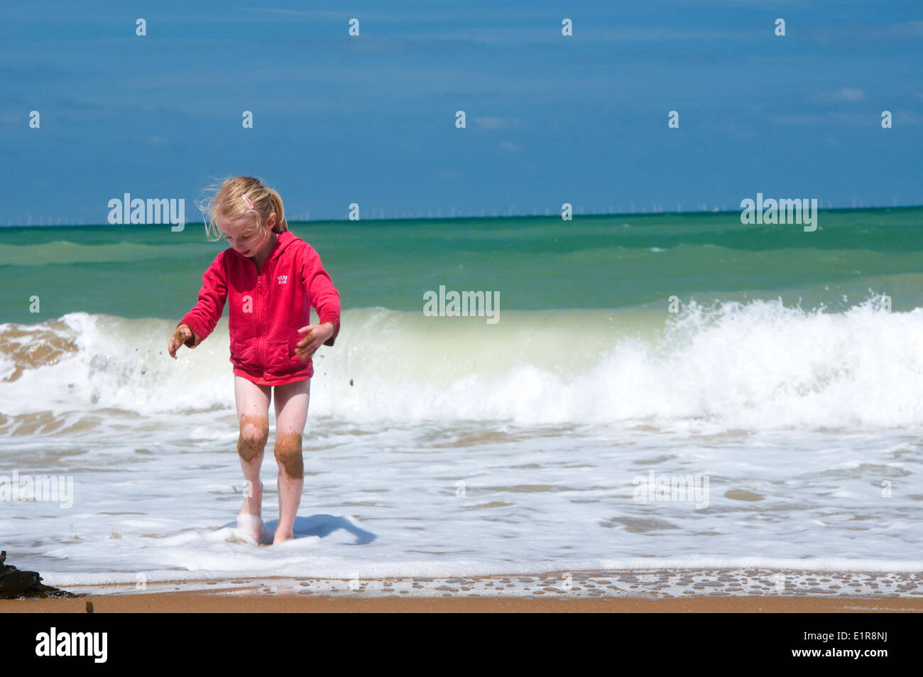 young girl playing with sand on the beach on a hot sunny day, with a rolling wave behind her. Stock Photo