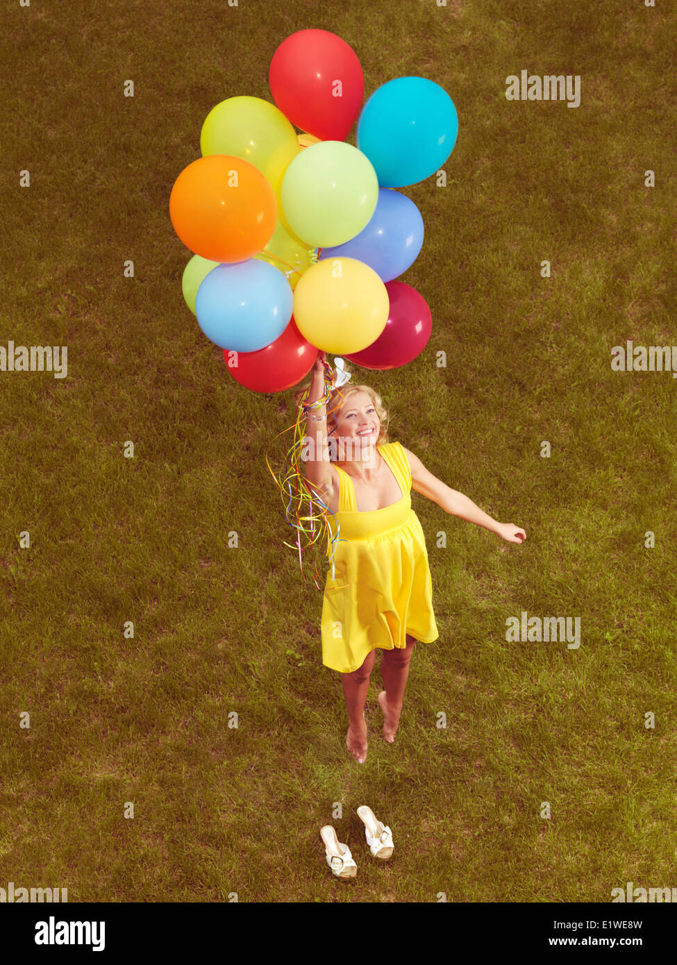 Happy young woman in yellow summer dress flying up from the ground with colorful helium balloons, retro stylized photo. Stock Photo