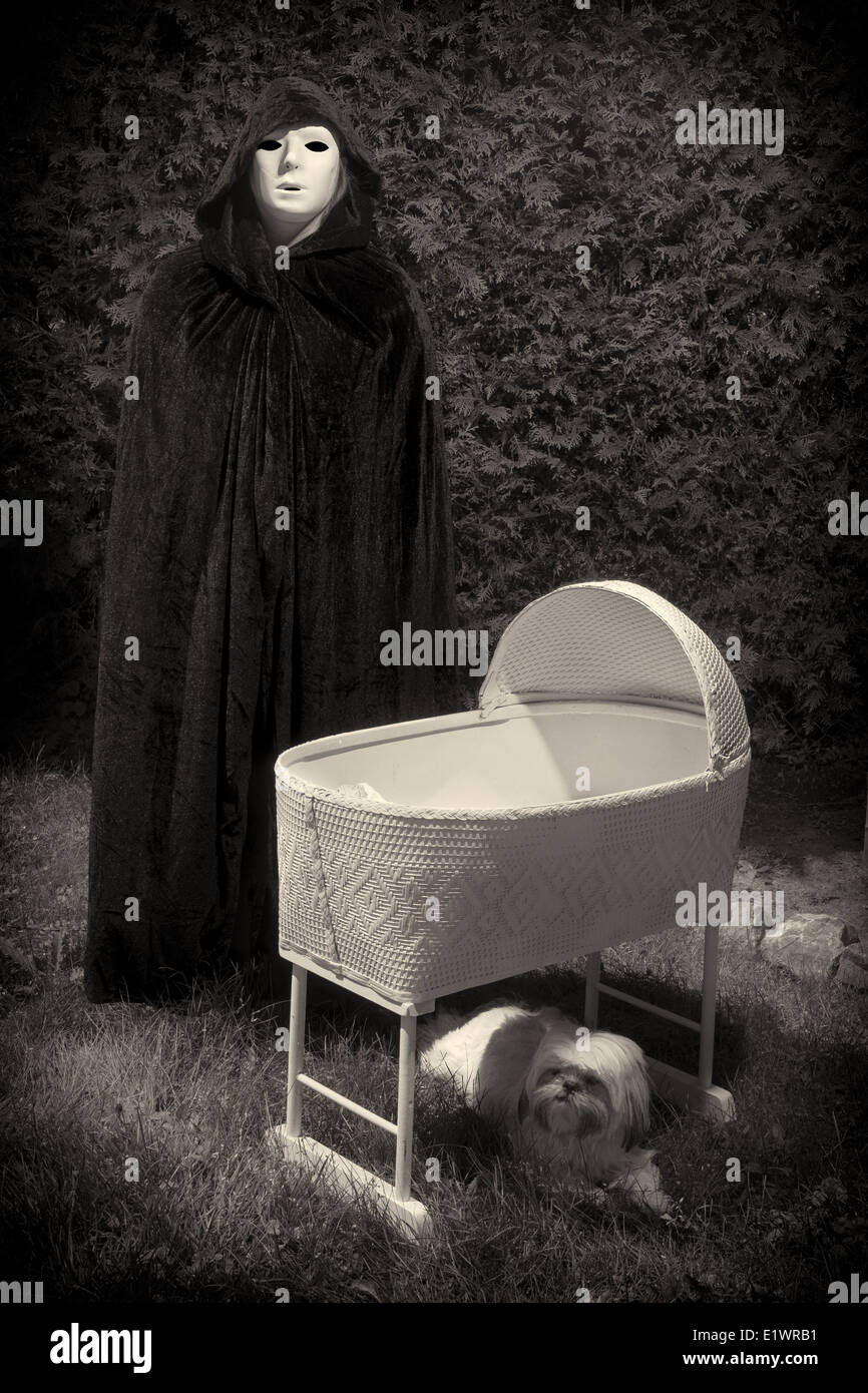 Cintage looking photo of a creepy masked and cloaked person standing next to an eerie, empty baby crib. Stock Photo