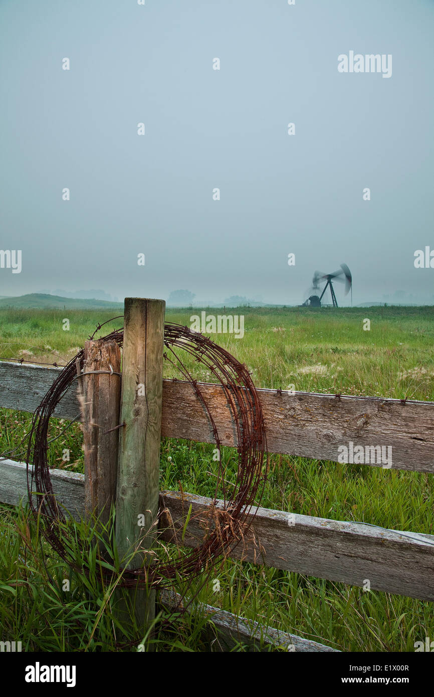 Barbed wire on fence post with pumpjack in background at dusk, Alberta, Canada Stock Photo