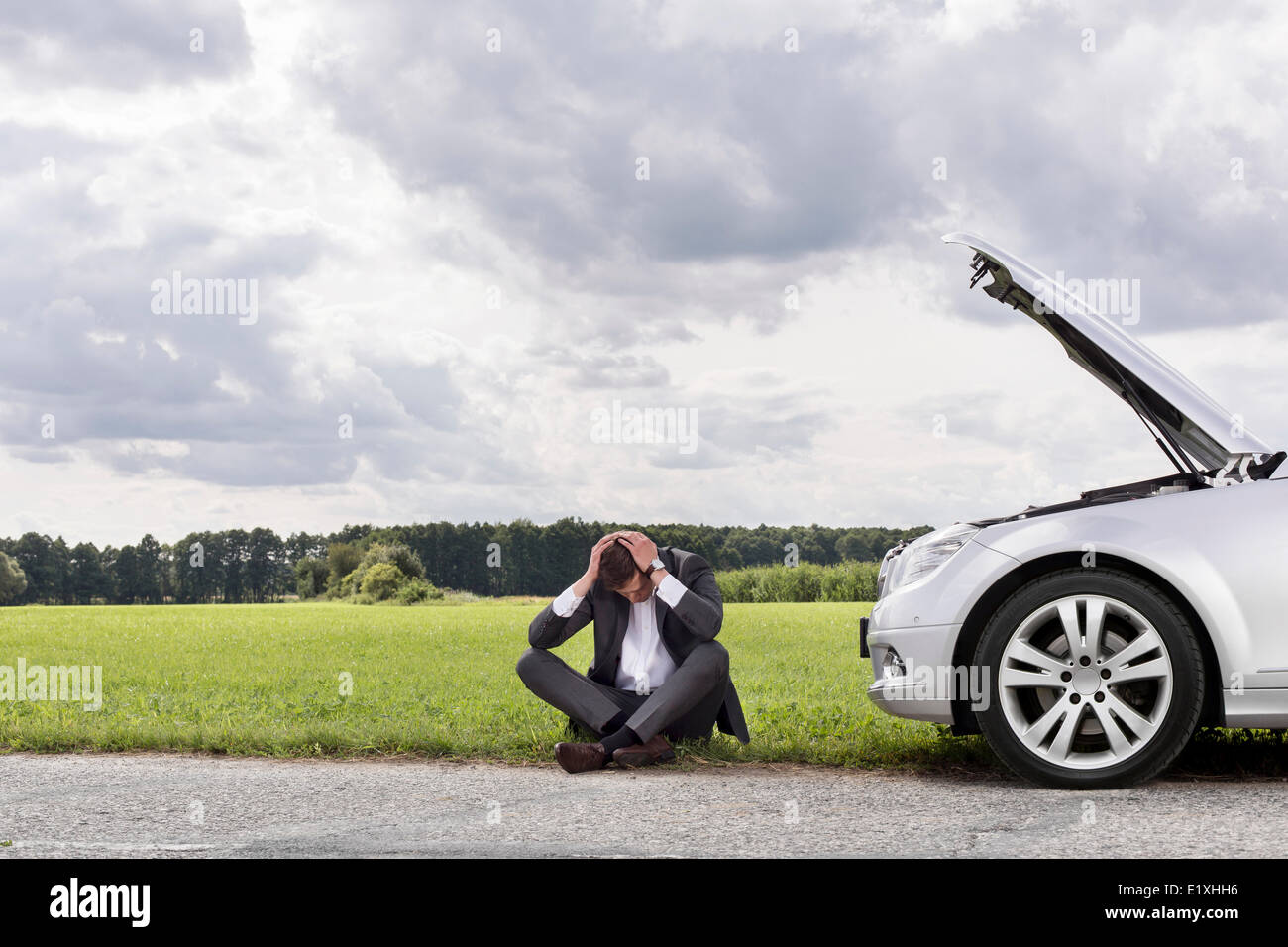 Full length of businessman with hands on head sitting by broken down car at countryside Stock Photo