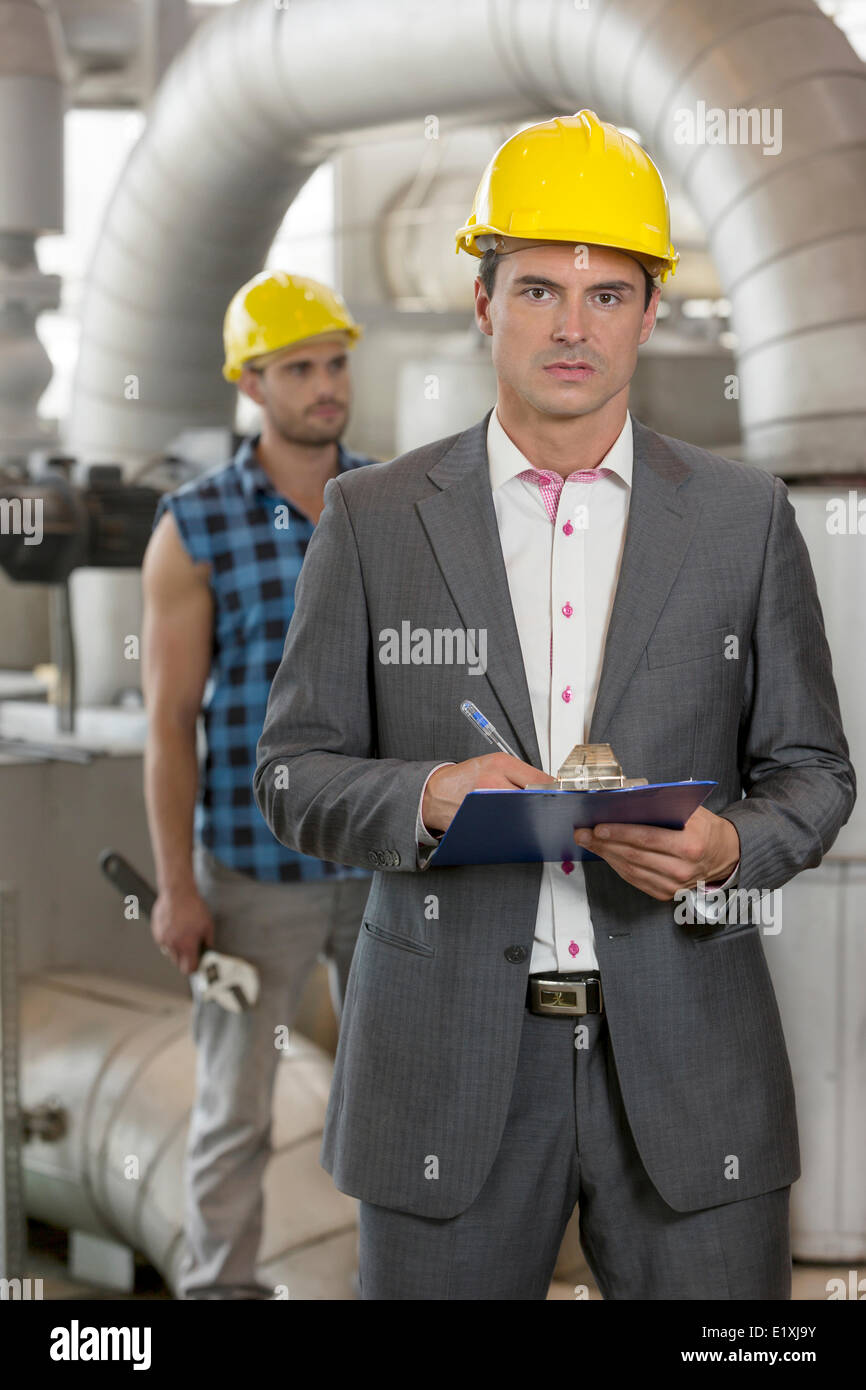 Portrait of young male manager writing on clipboard with manual worker in background at industry Stock Photo