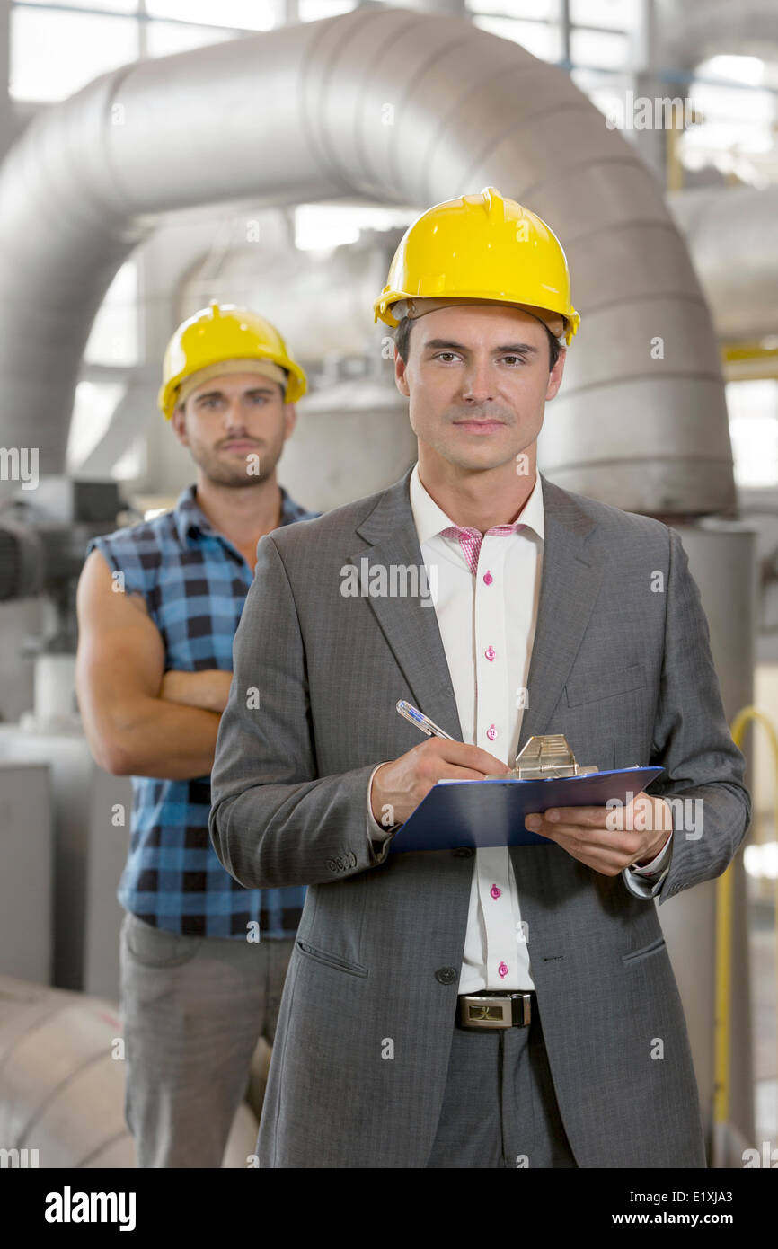 Portrait of confident young manager writing on clipboard with manual worker in background at industry Stock Photo