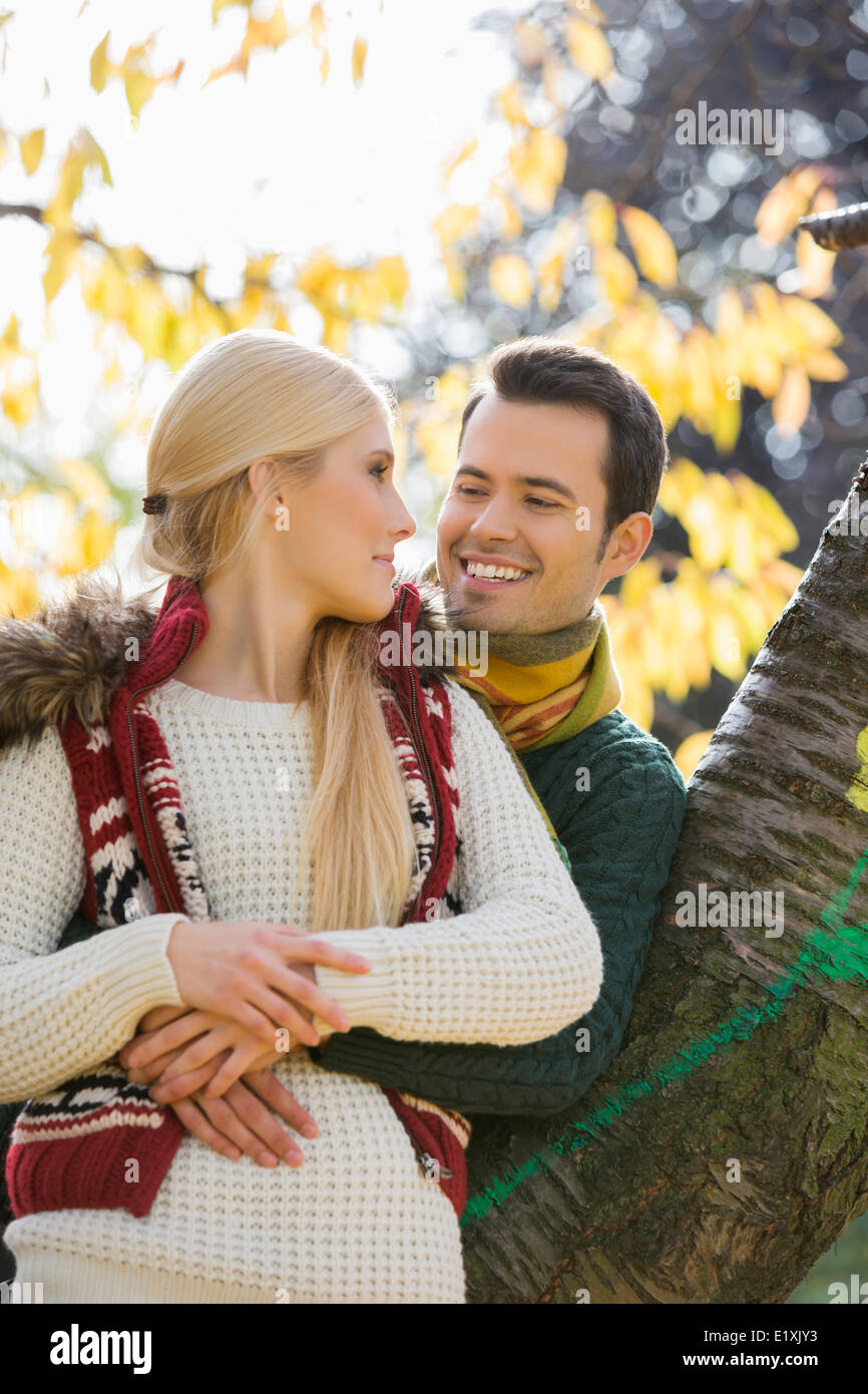 Happy young man hugging woman while leaning on tree trunk during autumn in park Stock Photo