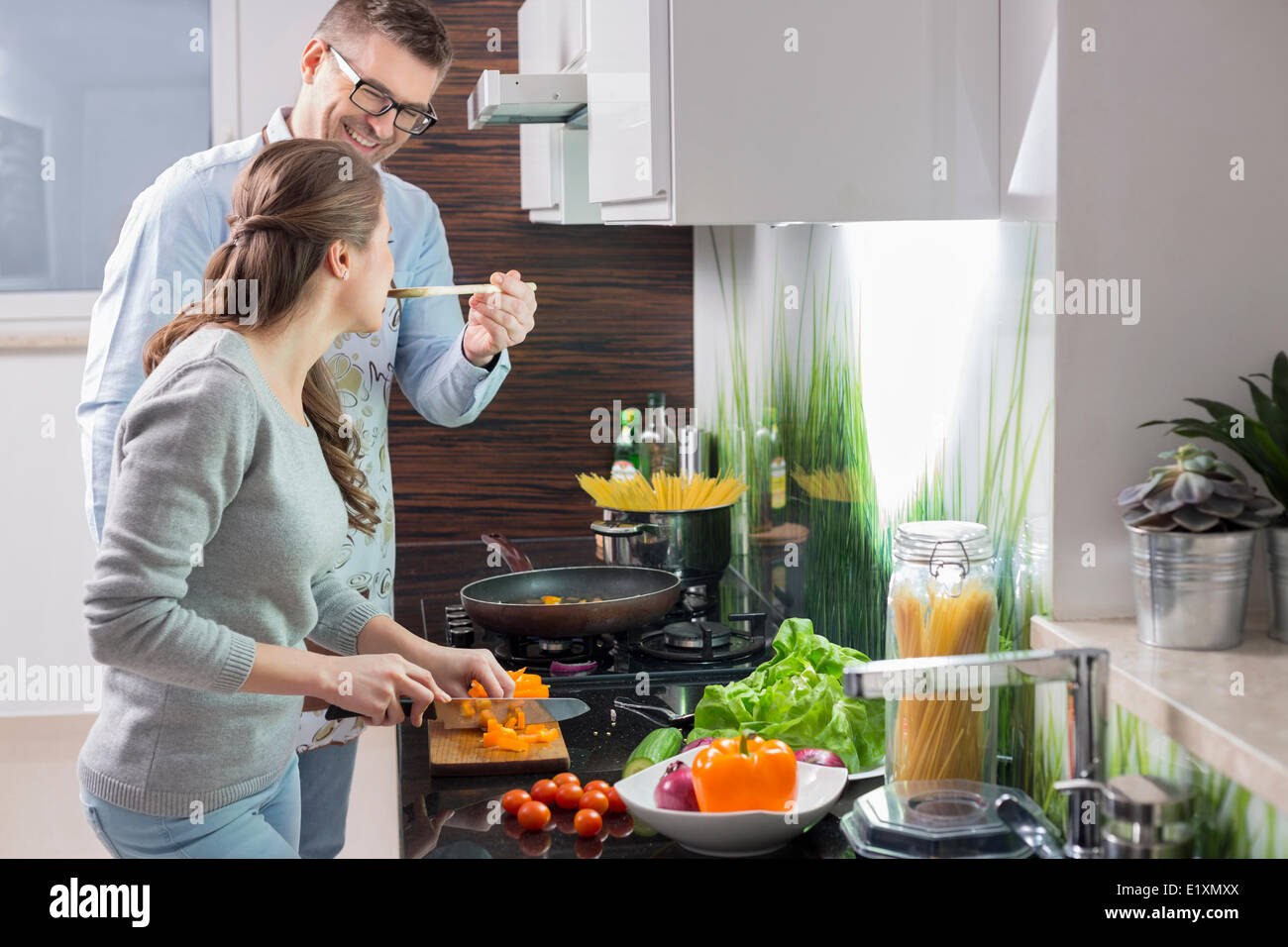 Happy man feeding food to woman cutting vegetables in kitchen Stock Photo