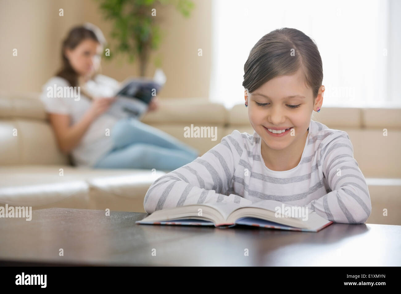 Smiling girl reading book with mother in background at home Stock Photo