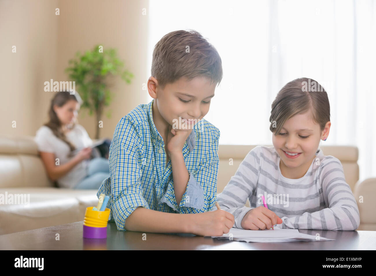 Siblings drawing together at table with mother in background Stock Photo