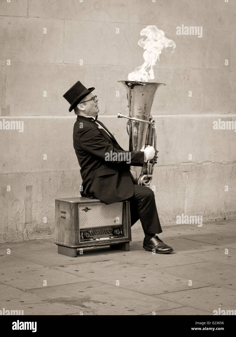 Busker, Christopher Werkowicz playing flaming tuba sitting on an old radio against a plain stone wall in sepia Stock Photo
