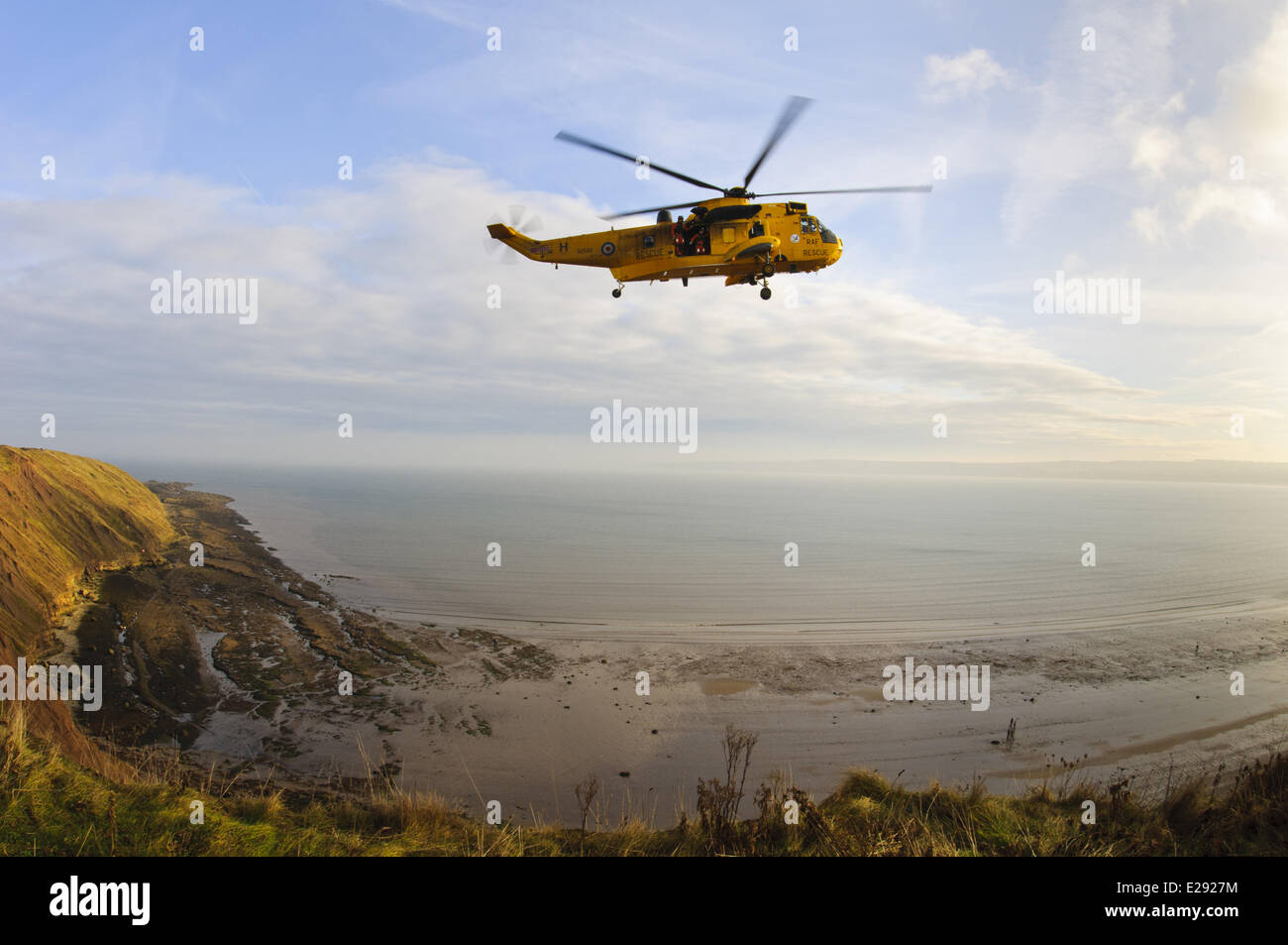 RAF Westland Sea King rescue helicopter with crew members standing in open doorway, in flight over rocky coastline and cliffs, looking from clifftop at Filey Country Park, Filey Brigg, North Yorkshire, England, November Stock Photo