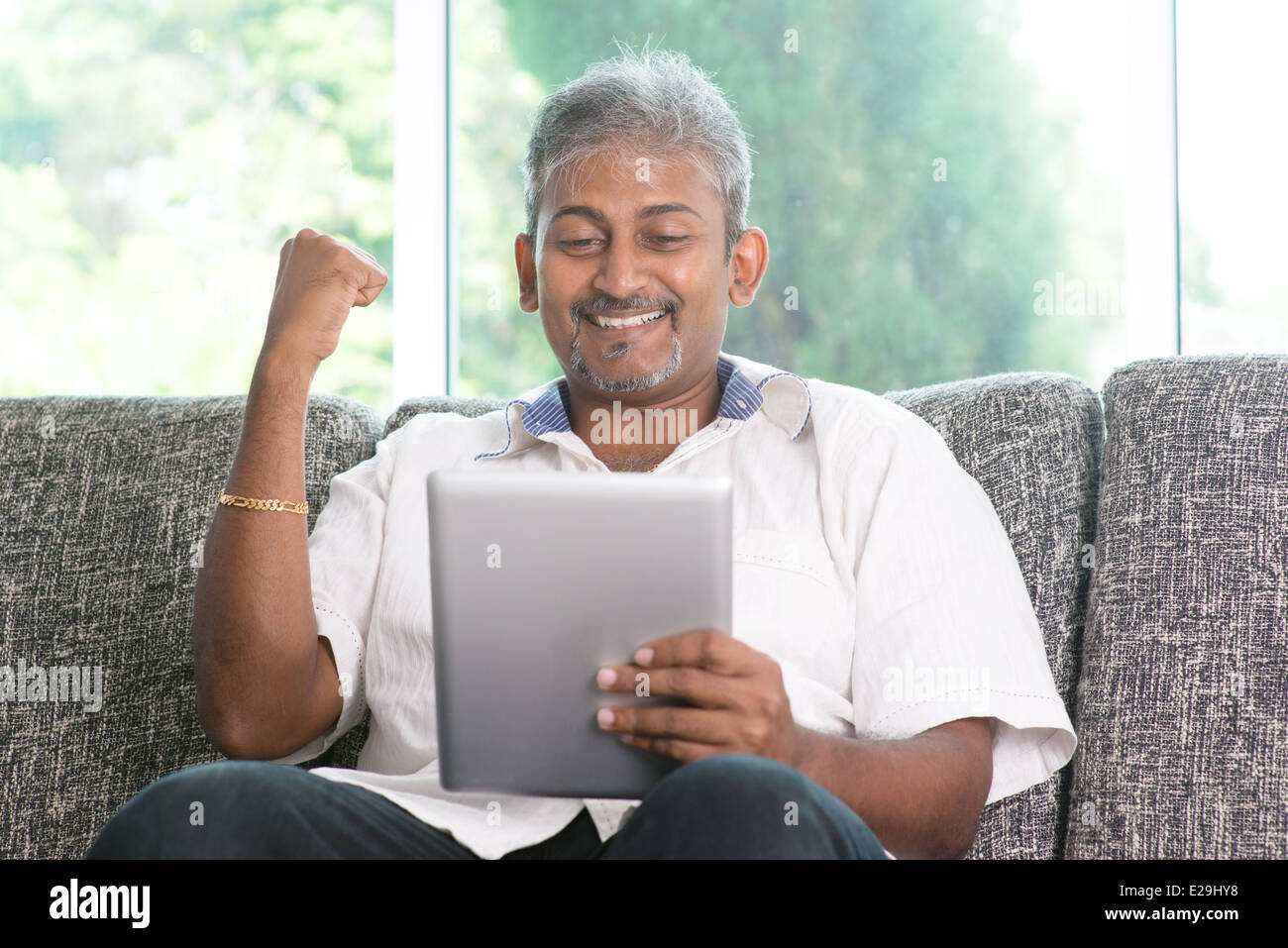 Portrait of middle aged Indian man reading on digital tablet computer and cheering at home. Stock Photo