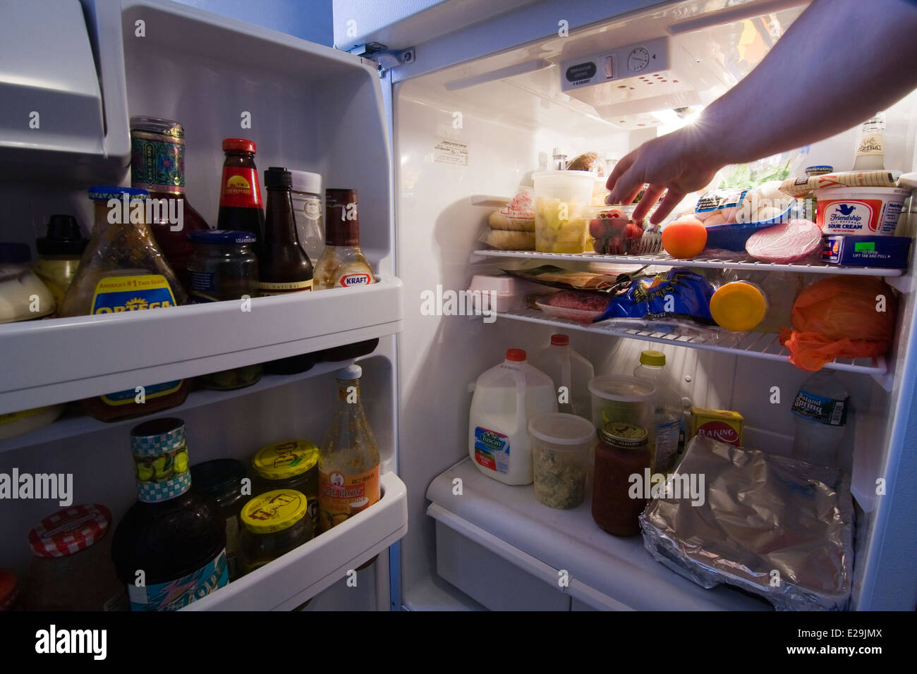 Door to a Refrigerator open with light on showing contents as a man's hand reaches for some fruit Stock Photo