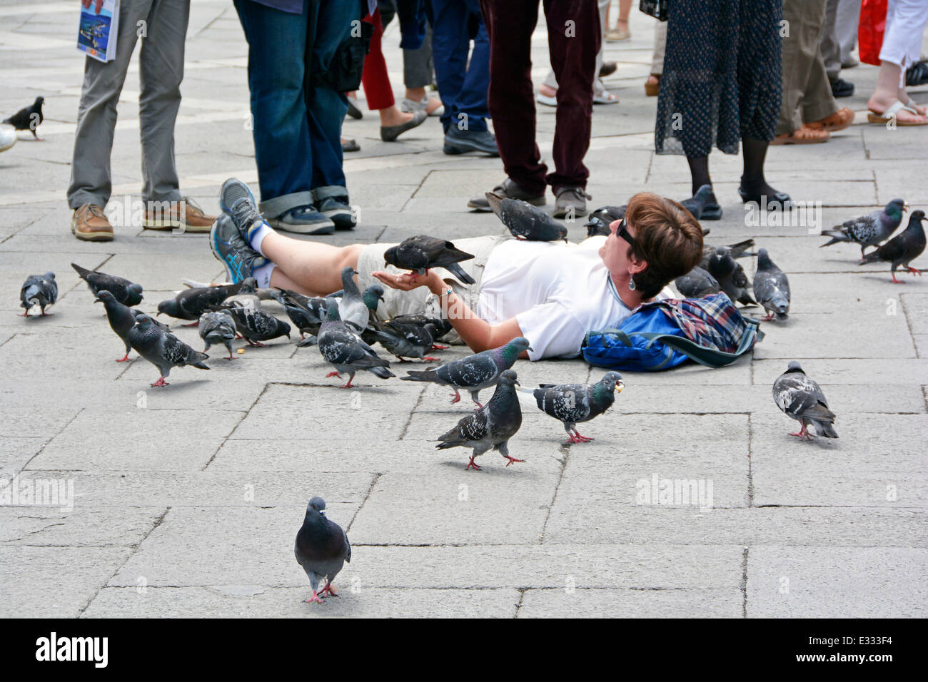 Woman laying down on paving slabs amongst the tourists in St Mark's Square Venice feeding the pigeons from her hands Stock Photo