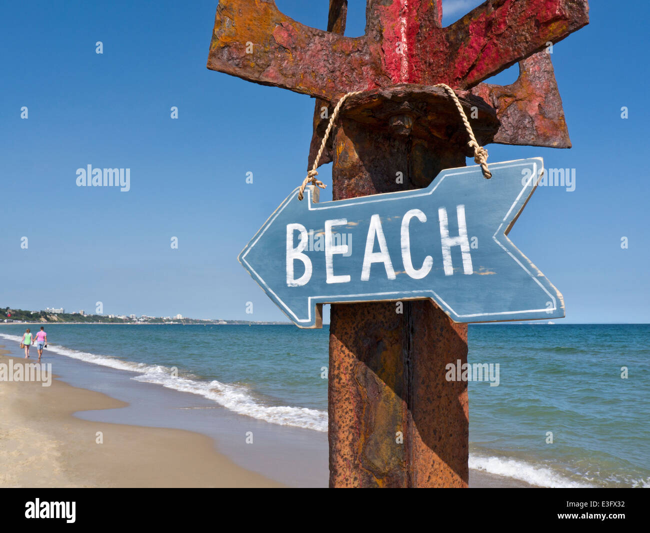 Wooden beach sign hanging on rustic metal post with couple in background strolling at waters edge Sandbanks Dorset UK Stock Photo