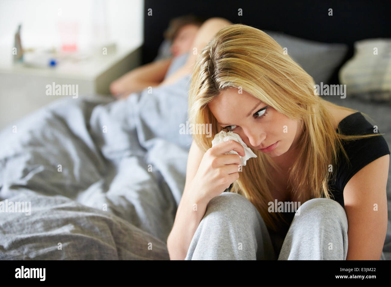 Sad Teenage Girl Sitting In Bedroom Whilst Boyfriend Sleeps Stock Photo