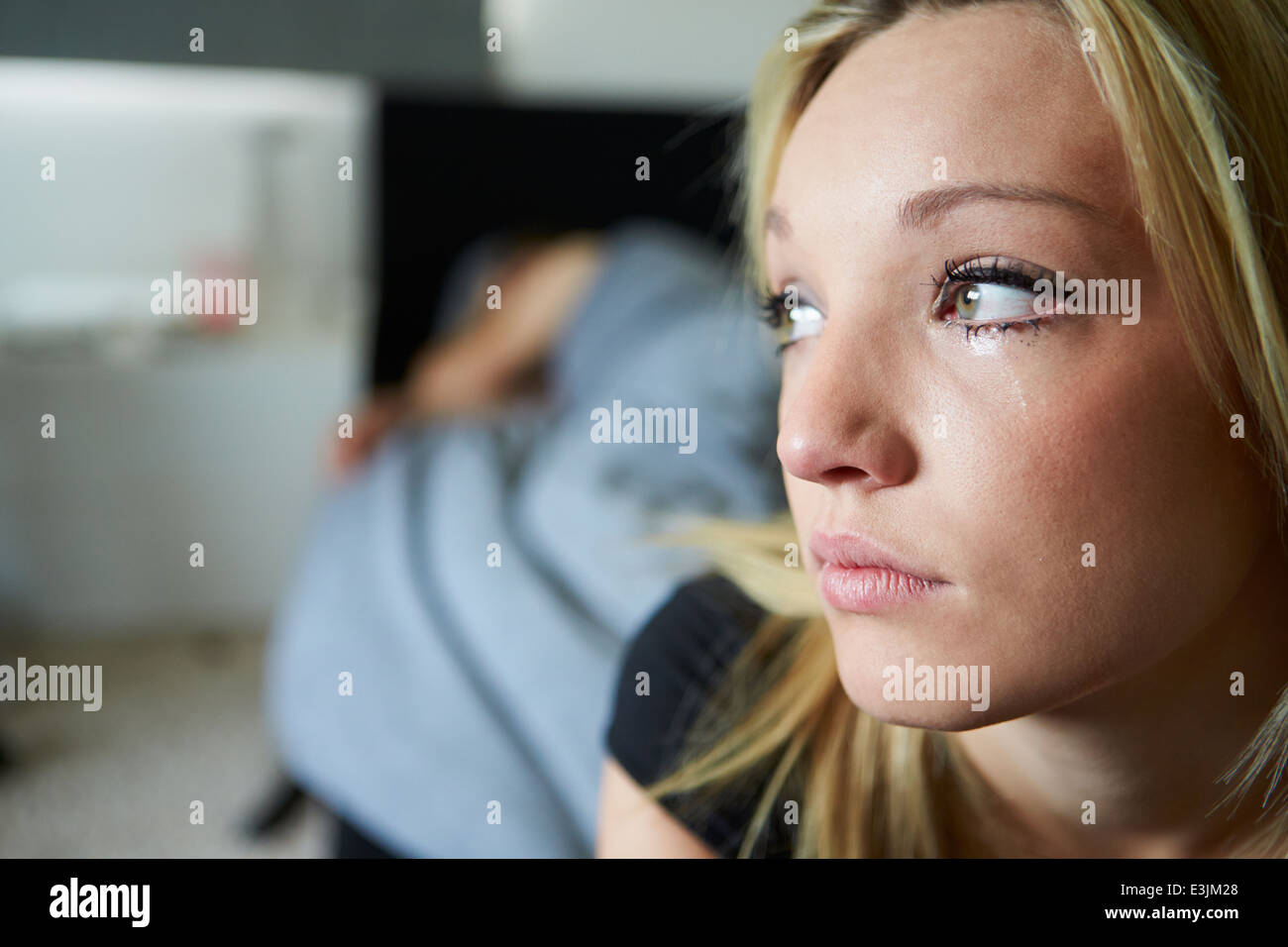 Sad Teenage Girl Sitting In Bedroom Whilst Boyfriend Sleeps Stock Photo