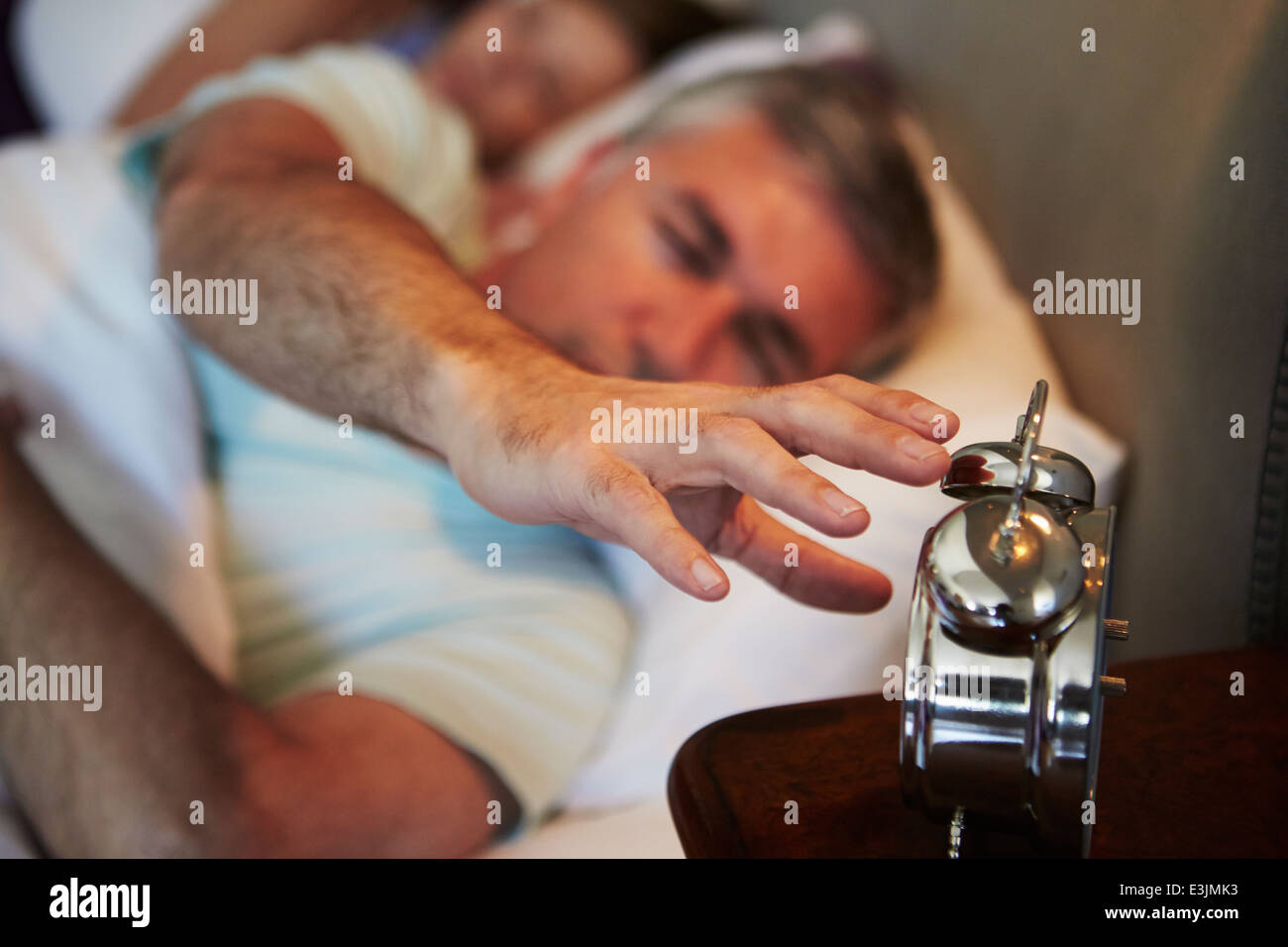 Couple In Bed With Man Reaching To Switch Off Alarm Clock Stock Photo