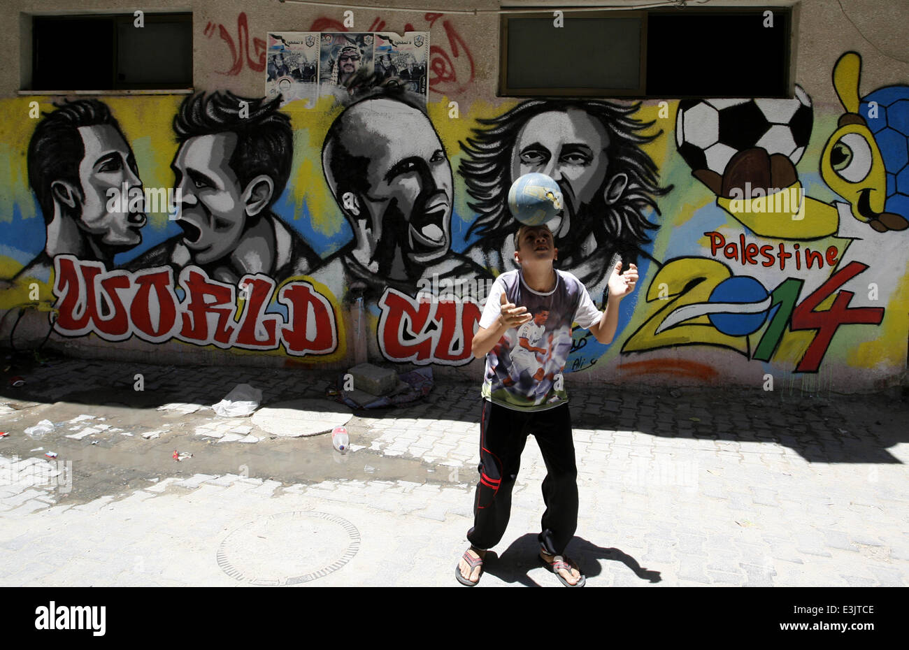 Gaza, Gaza Strip, Palestinian Territories. 23rd June, 2014. A Palestinian boy plays a ball in front of graffiti wall murals depicting football players the participants at 2014 World Cup Brazil (LtoR) Portugal's Cristiano Ronaldo, Argentina's Lionel Messi, Netherlands' Arjen Robben and Italy's Andrea Pirlo, in the Khan Yunis refugee camp in the southern Gaza Strip. on June 23, 2014. © Abed Rahim Khatib/NurPhoto/ZUMAPRESS.com/Alamy Live News Stock Photo