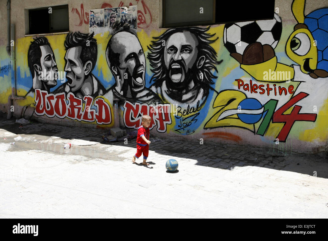 Gaza, Gaza Strip, Palestinian Territories. 23rd June, 2014. A Palestinian boy plays a ball in front of graffiti wall murals depicting football players the participants at 2014 World Cup Brazil (LtoR) Portugal's Cristiano Ronaldo, Argentina's Lionel Messi, Netherlands' Arjen Robben and Italy's Andrea Pirlo, in the Khan Yunis refugee camp in the southern Gaza Strip. on June 23, 2014. © Abed Rahim Khatib/NurPhoto/ZUMAPRESS.com/Alamy Live News Stock Photo