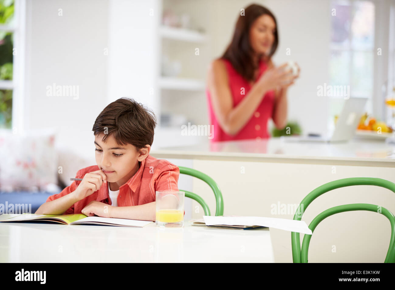 Son Does Homework As Mother uses Laptop In Background Stock Photo
