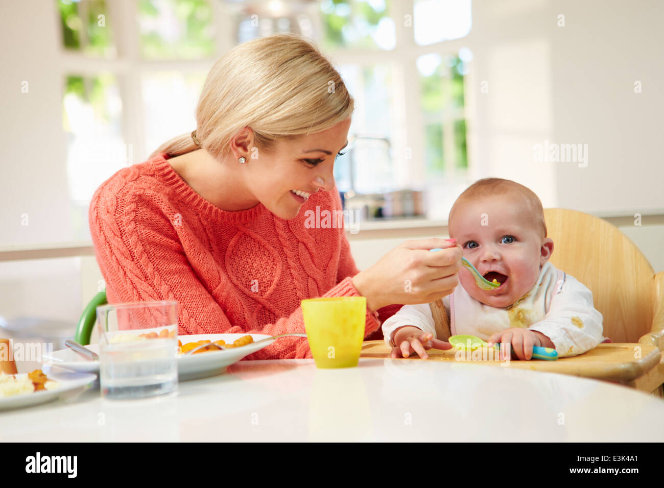 Mother Feeding Baby Sitting In High Chair At Mealtime Stock Photo