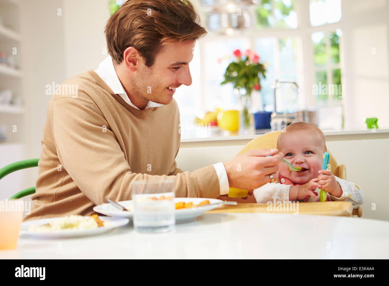 Father Feeding Baby Sitting In High Chair At Mealtime Stock Photo