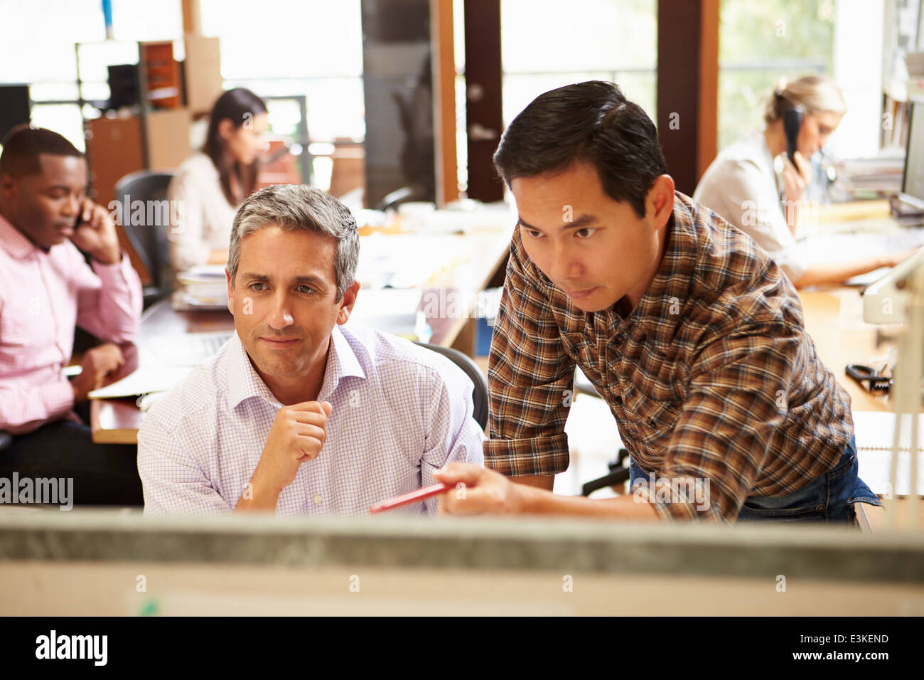 Two Colleagues Working At Desk With Meeting In Background Stock Photo