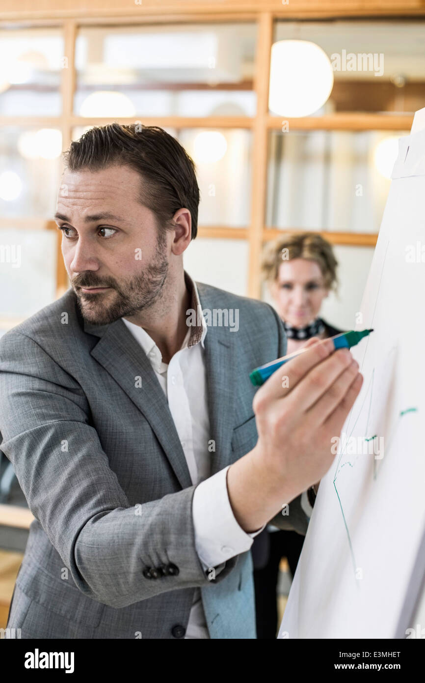 Mid adult businessman writing on flip chart with female colleague in background at office Stock Photo