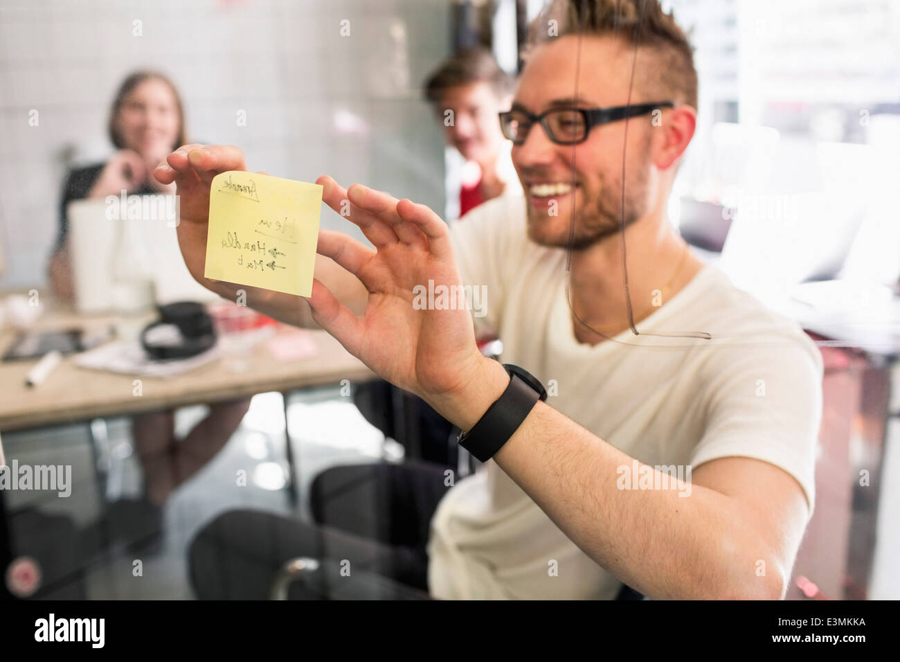 Young businessman sticking adhesive note on transparent glass with colleagues at background in new office Stock Photo
