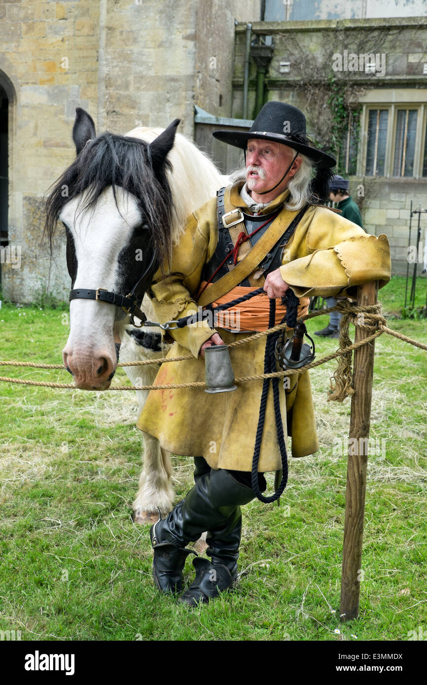 A lIving history reenactor portraying a cavalryman of English Civil war stood drinking beer, resting on a post with horse Stock Photo